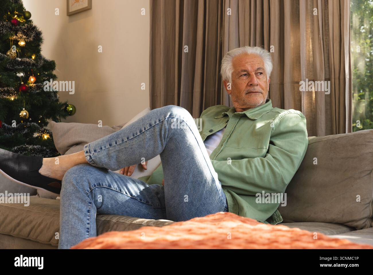 Ein älterer Mann sitzt im Wohnzimmer auf einem beigefarbenen Sofa, das mit Rostdecke neben dem Weihnachtsbaum bedeckt ist Stockfoto