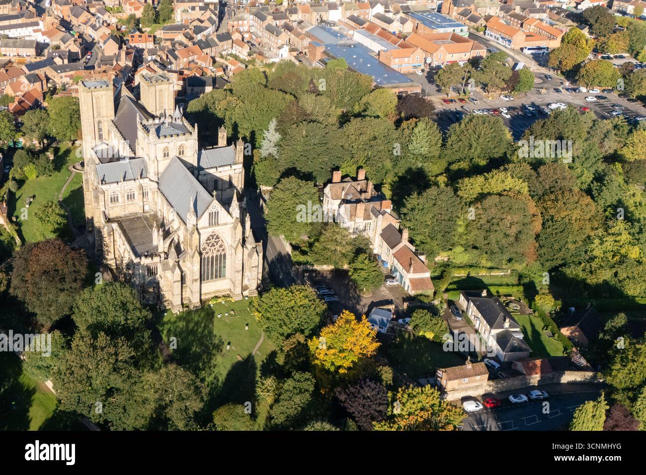 Luftaufnahme der Kathedrale von Ripon in Ripon, North Yorkshire, Großbritannien, aufgenommen bei klarem Spätsommerwetter mit hellblauem Himmel und warmem Sonnenlicht, das die beeindruckende gotische Architektur der Kathedrale unterstreicht. Das Bild zeigt die elegante frühe englische Westfront mit ihren Doppeltürmen, komplizierten Mauerwerken und bogenförmigen Fenstern, umgeben von den historischen Straßen der Stadt, nahe gelegenen Gebäuden, Dächern und Grünflächen einschließlich Parks und offenen Bereichen. Dieser Blick über der Drohne fängt die zentrale Position der Kathedrale in der kompakten mittelalterlichen Stadt ein und betont ihre Rolle als wichtiges Wahrzeichen des Kulturerbes Stockfoto