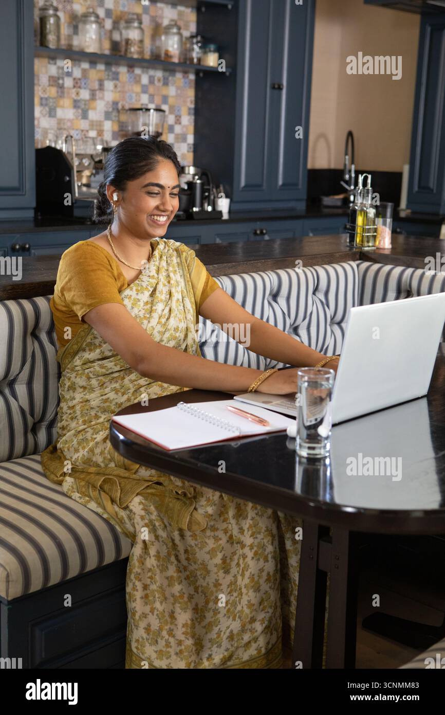Indianerin trägt gelben geblümten Sari, lächelt und tippt auf Laptop in der Cafeterküche, Kopierraum Stockfoto