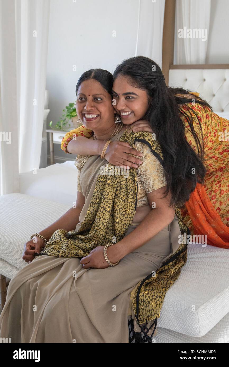 Indische Mutter und Tochter tragen traditionelle Kleidung, umarmen und lächeln im Schlafzimmer Stockfoto