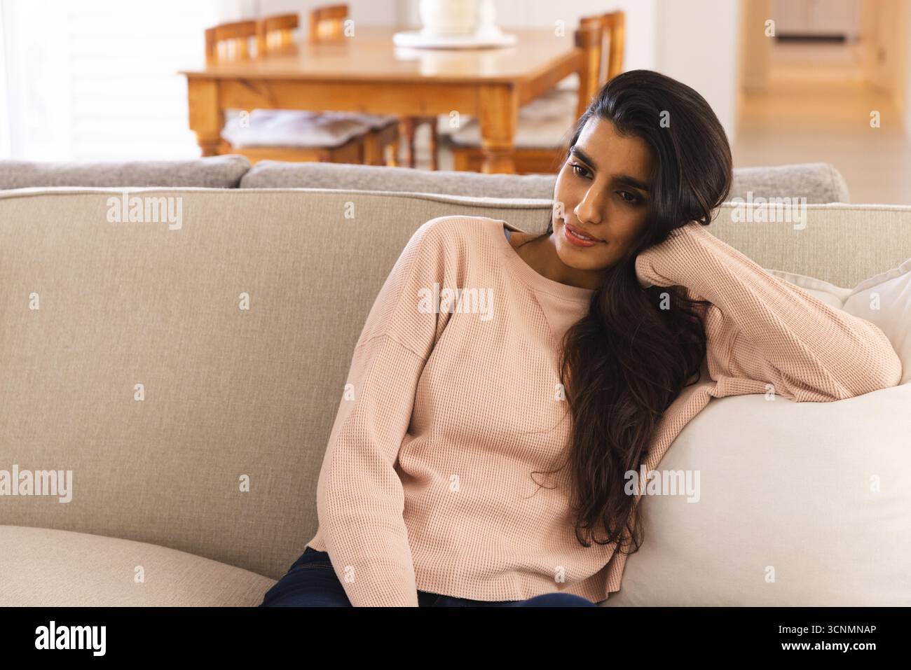 Asiatische Frau sitzt auf beigem Sofa im Wohnzimmer mit rosa Pullover und schaut zur Seite Stockfoto