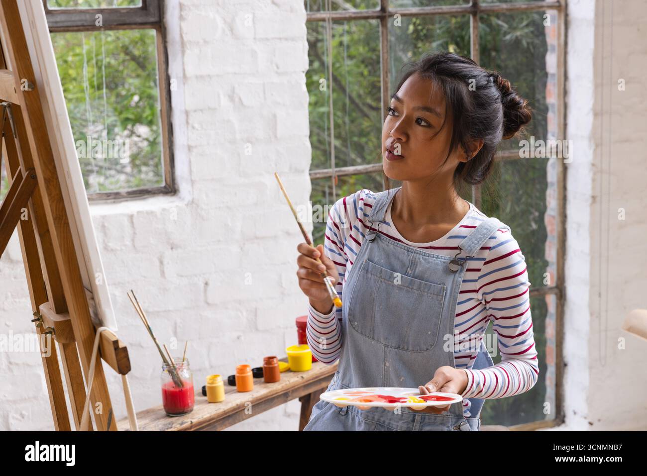Asiatische Malerin im Overall, die den nächsten Pinselstrich im Kunststudio mit Palette und Staffelei plant Stockfoto