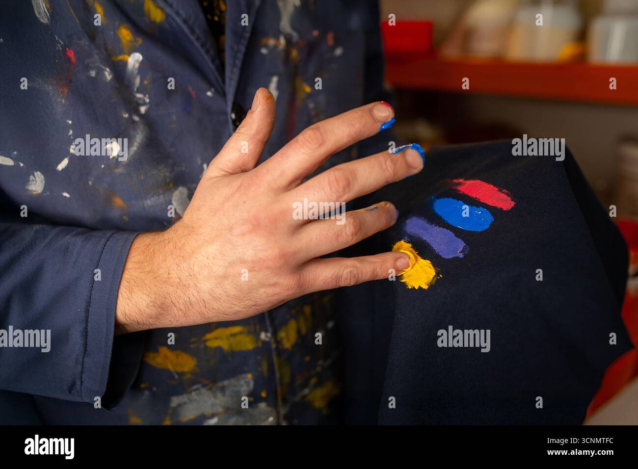 Handwerkstatt für Textildruck. Ein Handwerker mit schmutzigen Händen testet Siebdruckfarben auf einem Smock für die Bekleidungsherstellung. Stockfoto