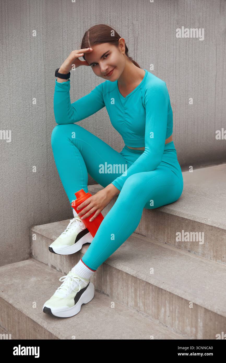 Fröhliche Frau in Mode sportliches Outfit im Freien mit Wasserflasche und lächelndem Lächeln. Fitness-Frau, die nach dem Training eine Pause macht Stockfoto