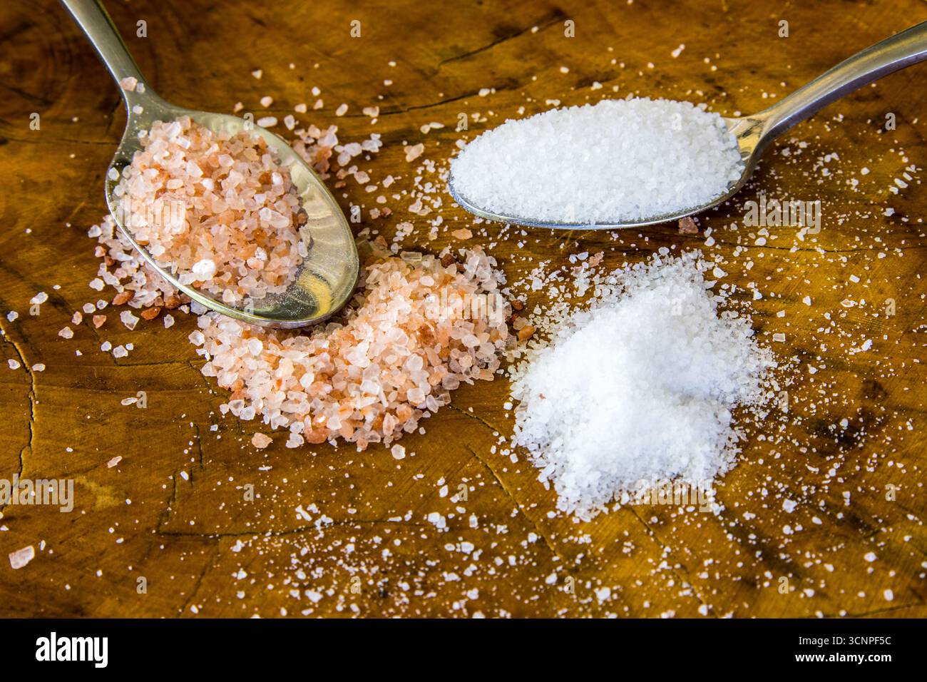 Zwei Teelöffel pinkfarbenes Himalaya-Salz und weißes Tafelsalz, umgeben von verschüttetem Salz Stockfoto