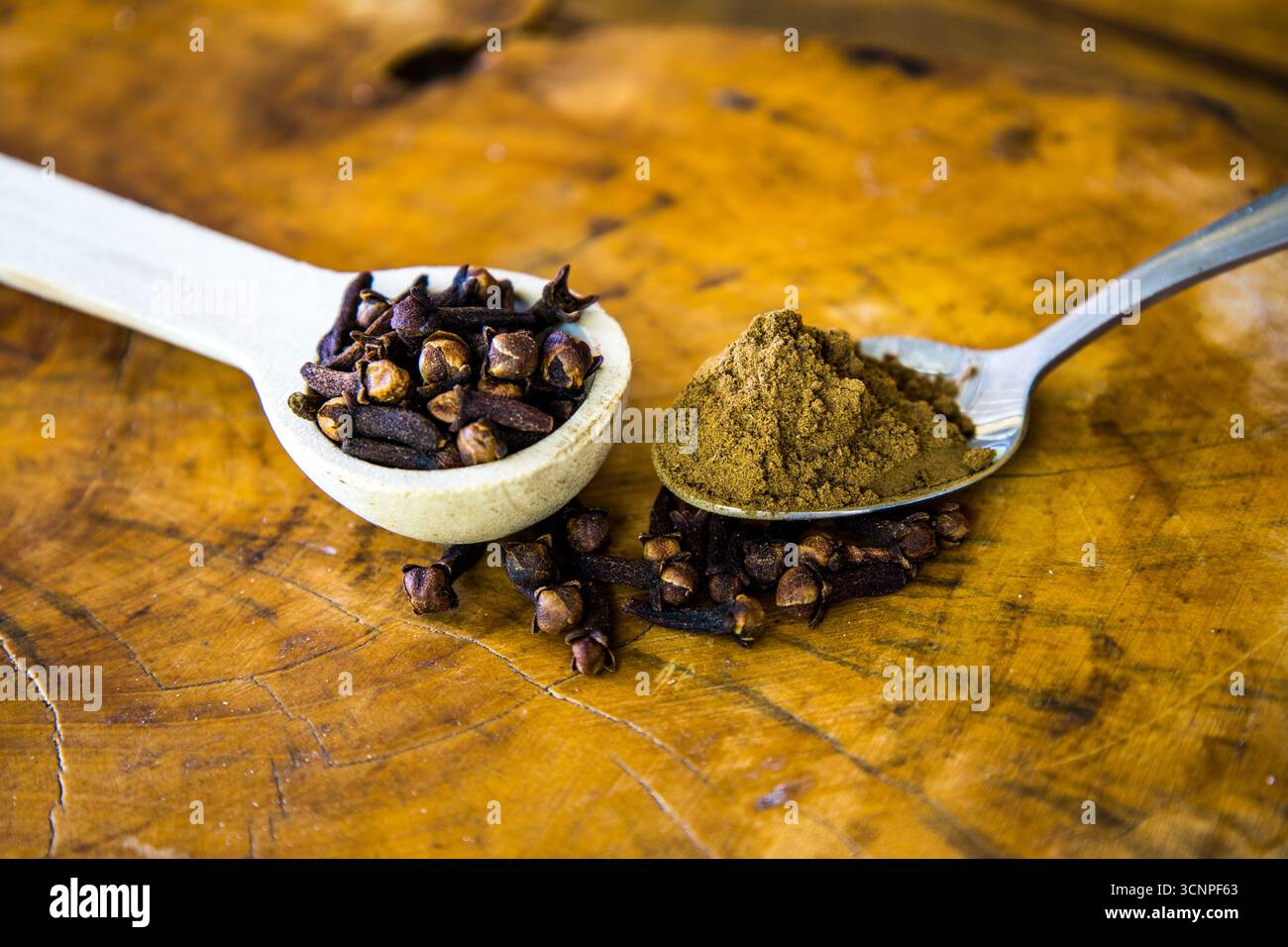 Zwei verschiedene Teelöffel, gefüllt mit gemahlenen Zehen und ganzen Zehen und umgeben von verschütteten Gewürzen auf hölzernem Hintergrund. Stockfoto