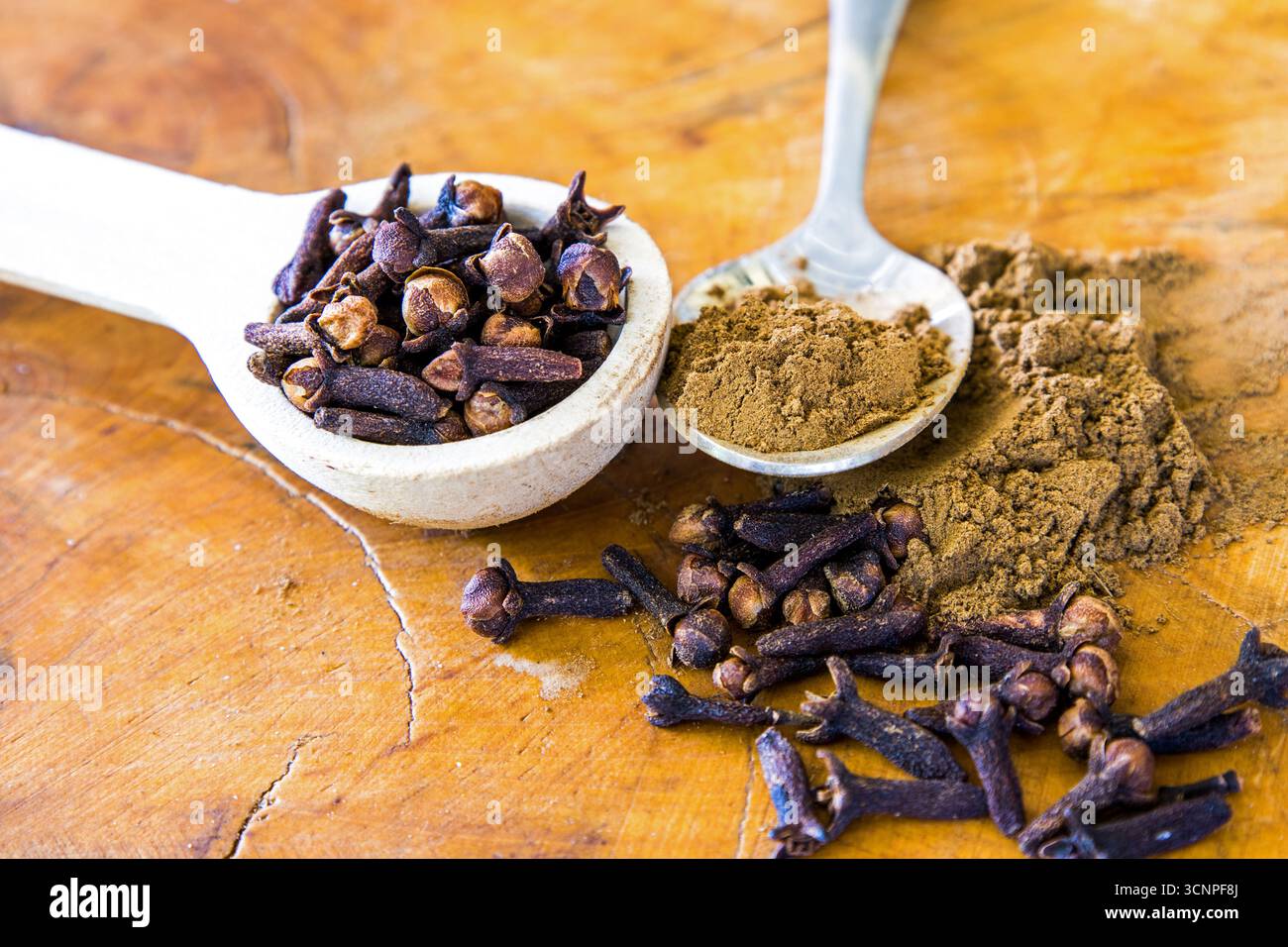 Zwei verschiedene Teelöffel, gefüllt mit gemahlenen Zehen und ganzen Zehen und umgeben von verschütteten Gewürzen auf hölzernem Hintergrund. Stockfoto