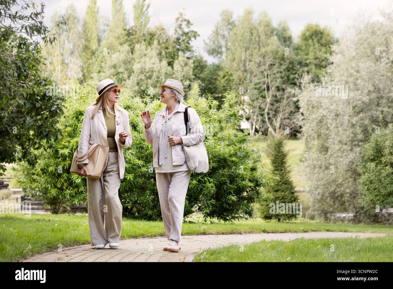 Porträt in voller Länge von zwei älteren Frauen, die in stilvollen, hellbeigen und cremefarbenen Casual-Outfits nach draußen gehen, mit Hüten, Leinenhosen, Jacken, Tasche und Stockfoto