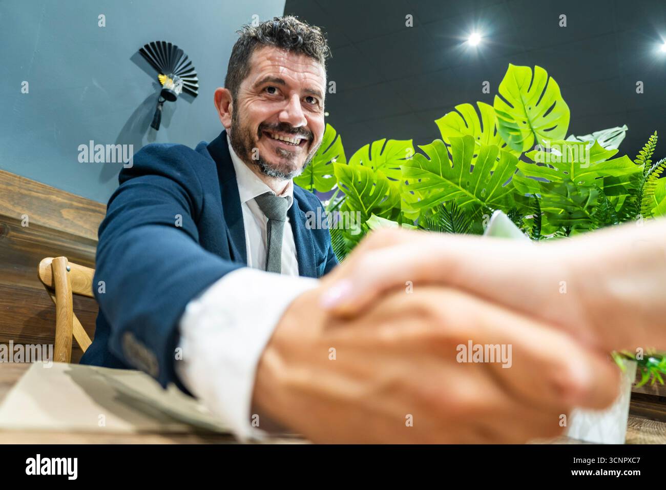 Ein selbstbewusster Geschäftsmann im Anzug lächelt warm und schüttelt die Hände über einen Schreibtisch Stockfoto