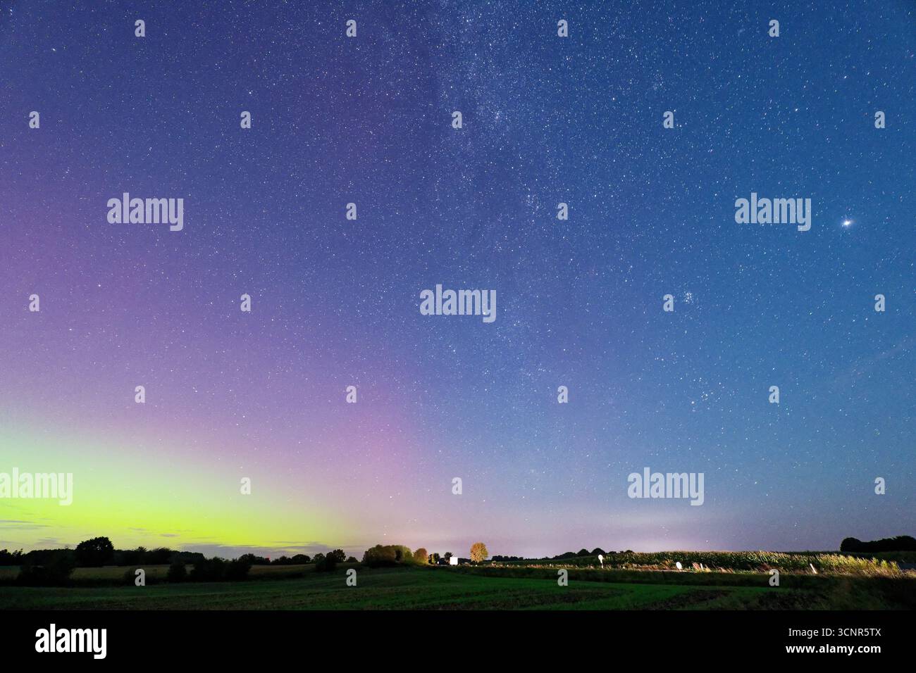 aurorafarben welken über einem weiten Feld, während weit entfernte Autos helle Scheinwerferlinien am Horizont und gelb-weiß-rosa-violette und blaue Schichten hinterlassen Stockfoto