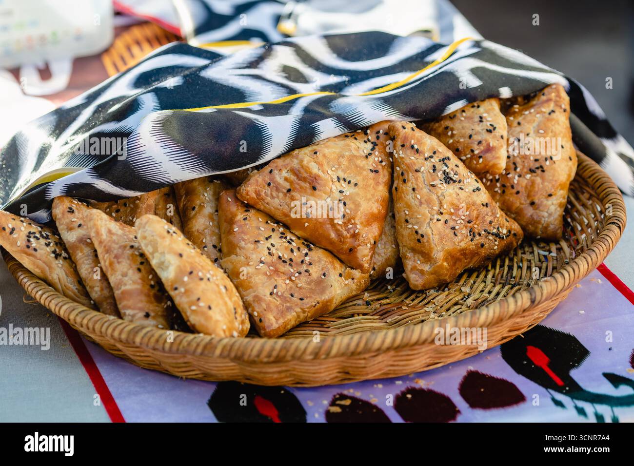 Korb mit frisch gebackenem usbekischen samsa-Gebäck, bedeckt mit einem dekorativen Tuch. Traditioneller zentralasiatischer Snack mit knusprigem Teig, Fleisch und Gewürzen Stockfoto