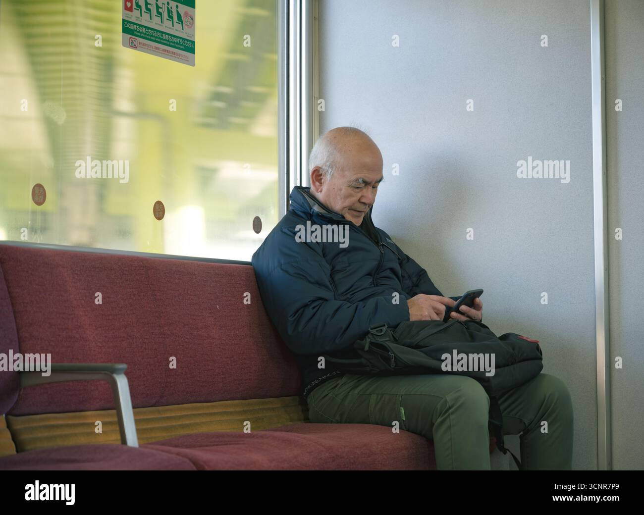 Ein älterer Mann in einer marineblauen Jacke sitzt auf einer kastanienbraunen japanischen Zugbank, in sein Handy eingesogen Stockfoto