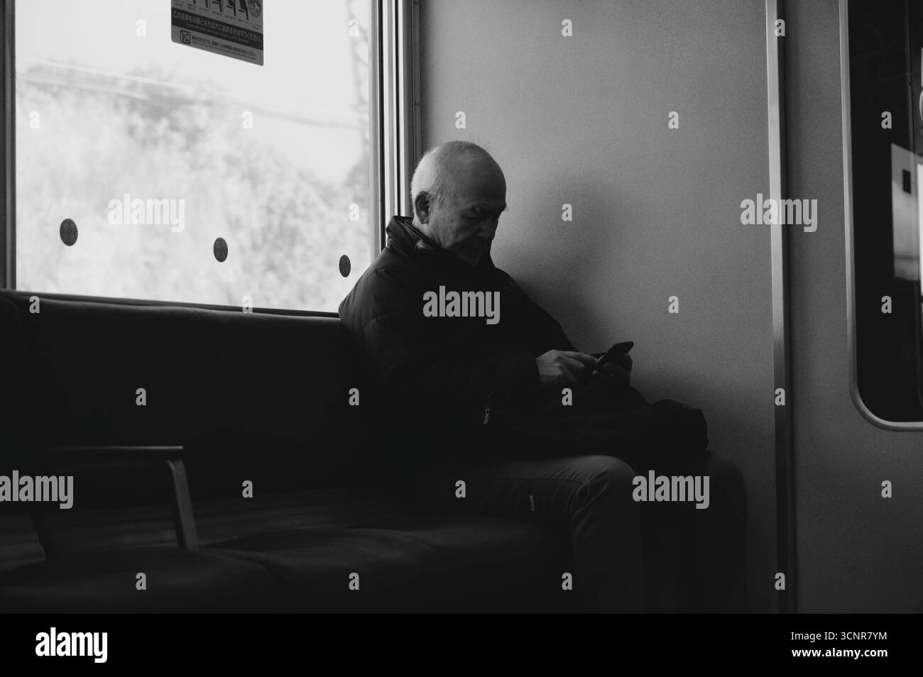 Ein älterer Mann sitzt am Fenster eines Nara-Zuges, in sein Handy eingesaugt, in Schwarz-weiß gefangen Stockfoto
