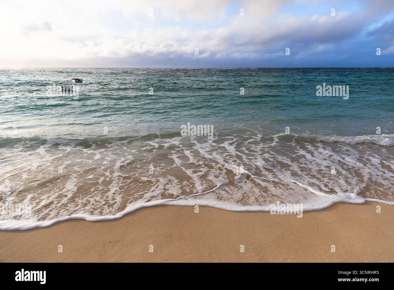 Ein ruhiger Blick auf die Küste mit sanften Wellen, die unter einem dramatischen Himmel auf die Sandküste strömen. Kleine Boote schweben auf türkisfarbenem Wasser, während der Horizont lockt Stockfoto