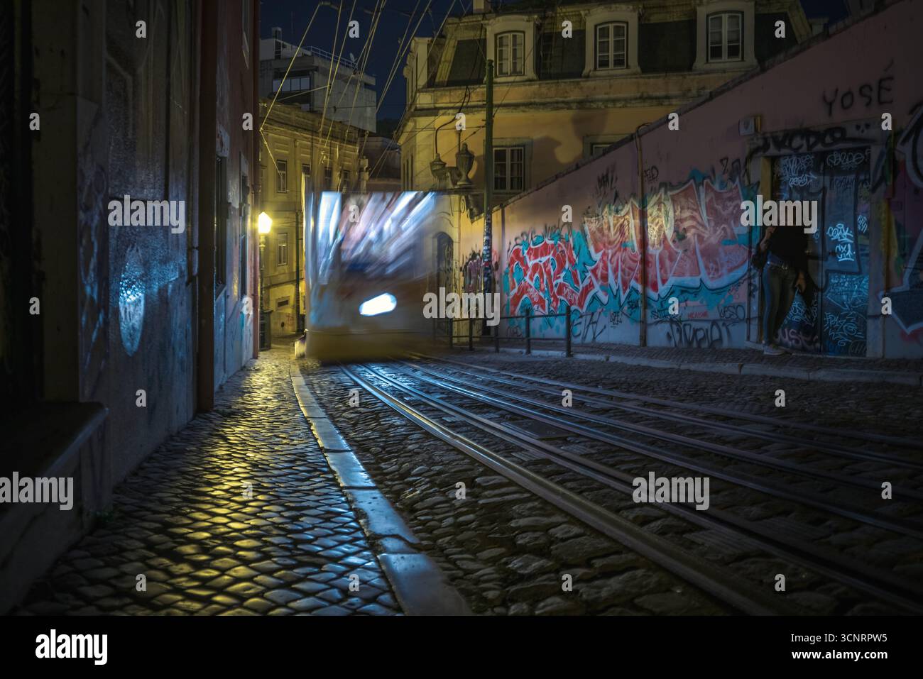 Nächtliche Straßenbahnfahrt mit Graffiti-Wänden und Kopfsteinpflasterstraße in Lissabon Portugal, städtische Straßenfotografie mit Lichtspuren Stockfoto