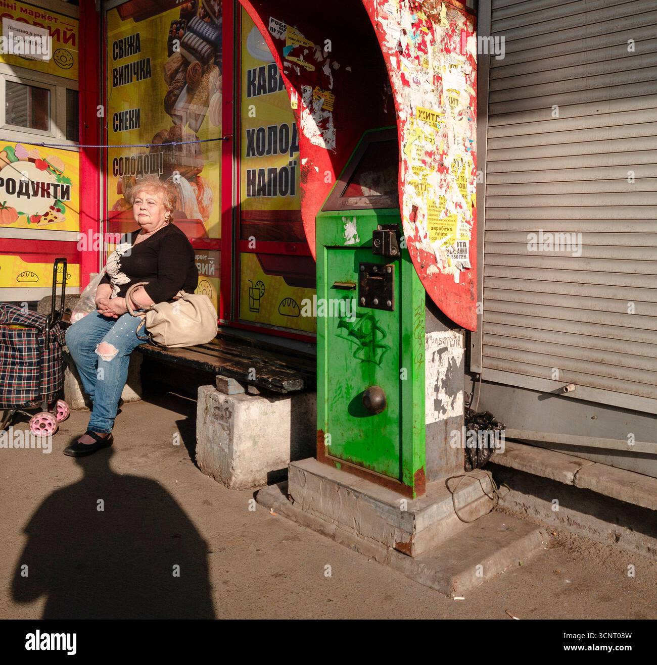 Kiew, Ukraine 5. Mai 2025, ältere Frau, die auf einer Bank in der Nähe des Kiosks mit bunten Plakaten und alten Verkaufsautomaten im Nachmittagssonnenlicht auf einer Straße in Kiew Stadt Ukraine sitzt Stockfoto