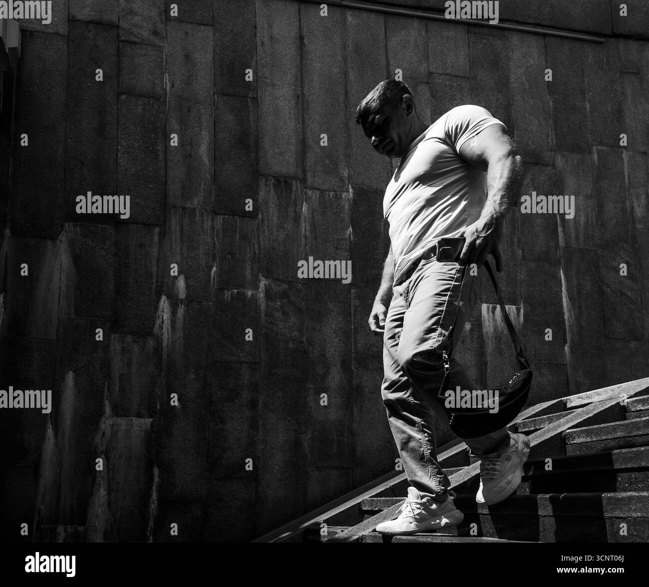 Kiew, Ukraine 3. Juni 2025, muskulöser Mann steigt in Kiew eine Betontreppe hinab, trägt eine kleine Tasche, die in Schwarz-weiß mit einem auffälligen Lichtkontrast gefangen ist und Stärke, Einsamkeit und Bewegung symbolisiert Stockfoto