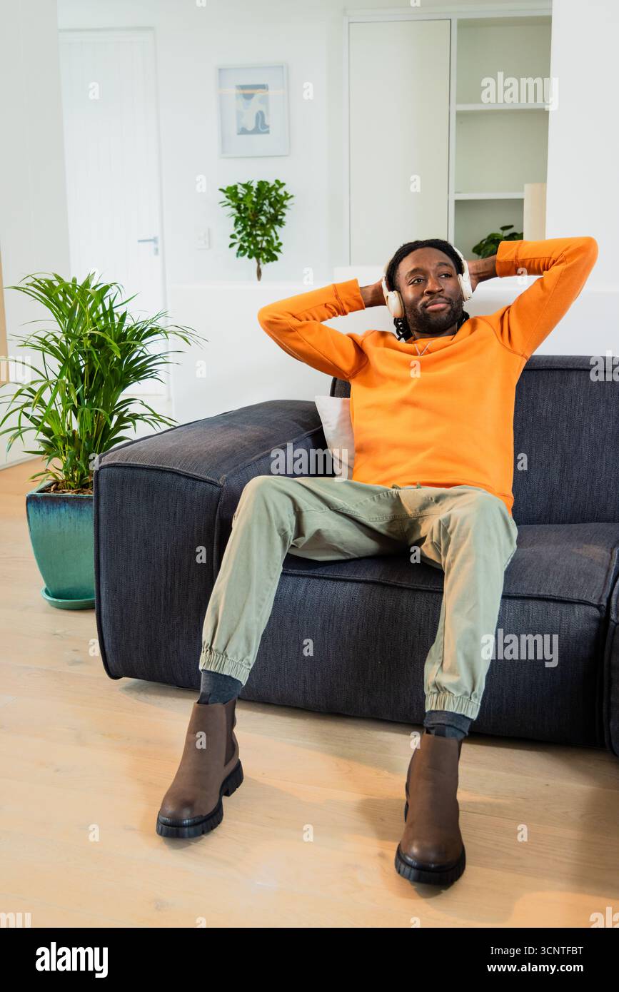 Afroamerikanischer Erwachsener, der sich mit kabellosen Kopfhörern auf dem dunkelgrauen Sofa im Wohnzimmer zurücklehnt Stockfoto