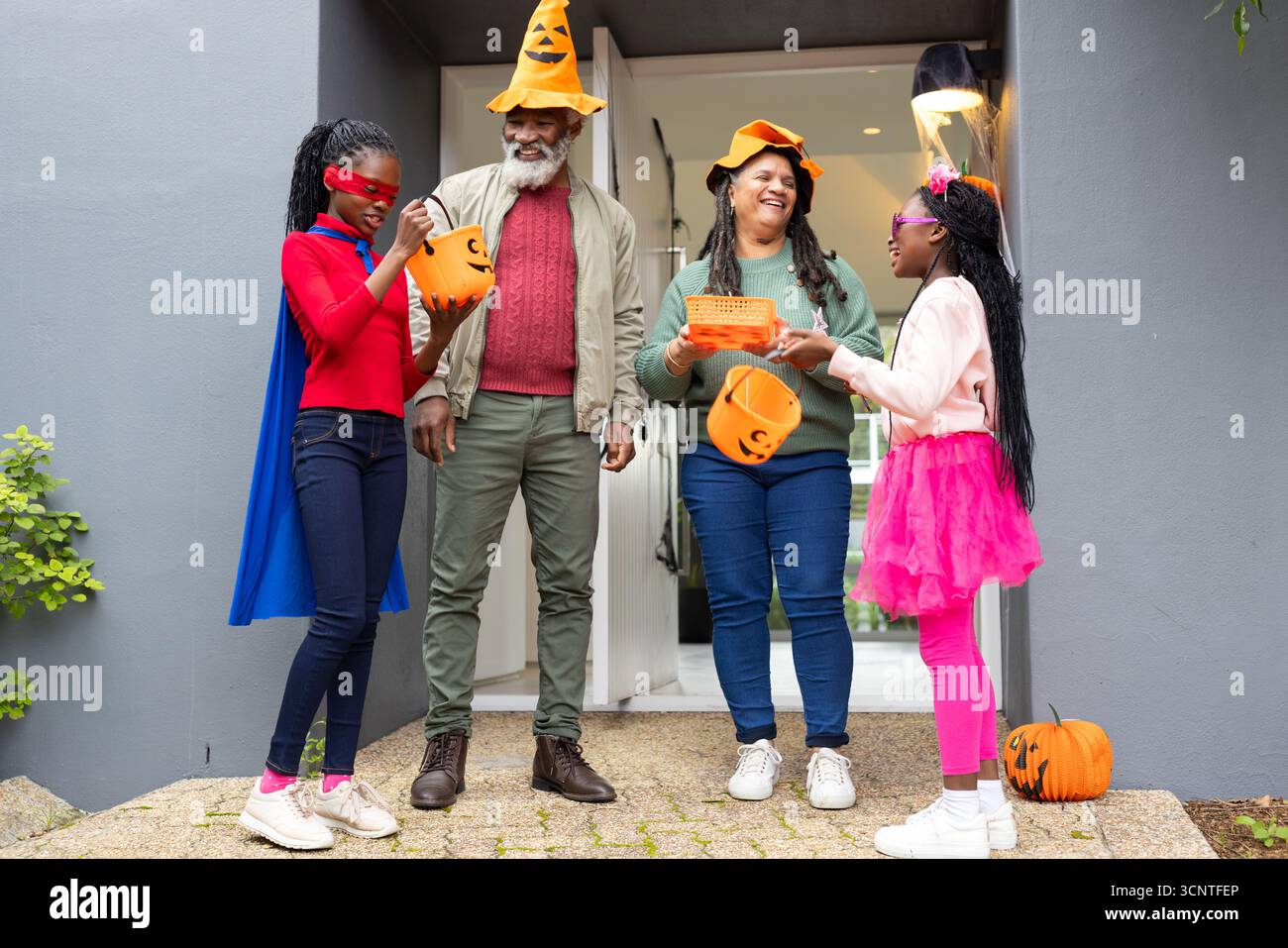 Diverse Seniorenpaare mit Enkelkindern, die auf der Veranda in Kürbishüten posieren und Kürbiseimer halten Stockfoto