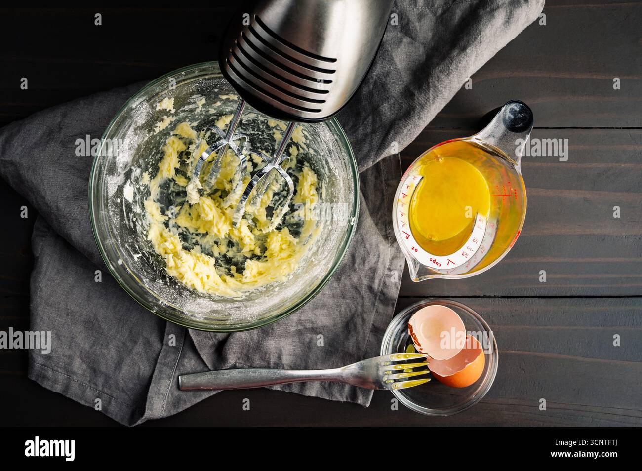Rahmbutter und Puderzucker in Glasrührschüssel: Geschlagene Butter und Süßwarenzucker mit Handrührgerät, geschlagenem Ei und einer Gabel Stockfoto