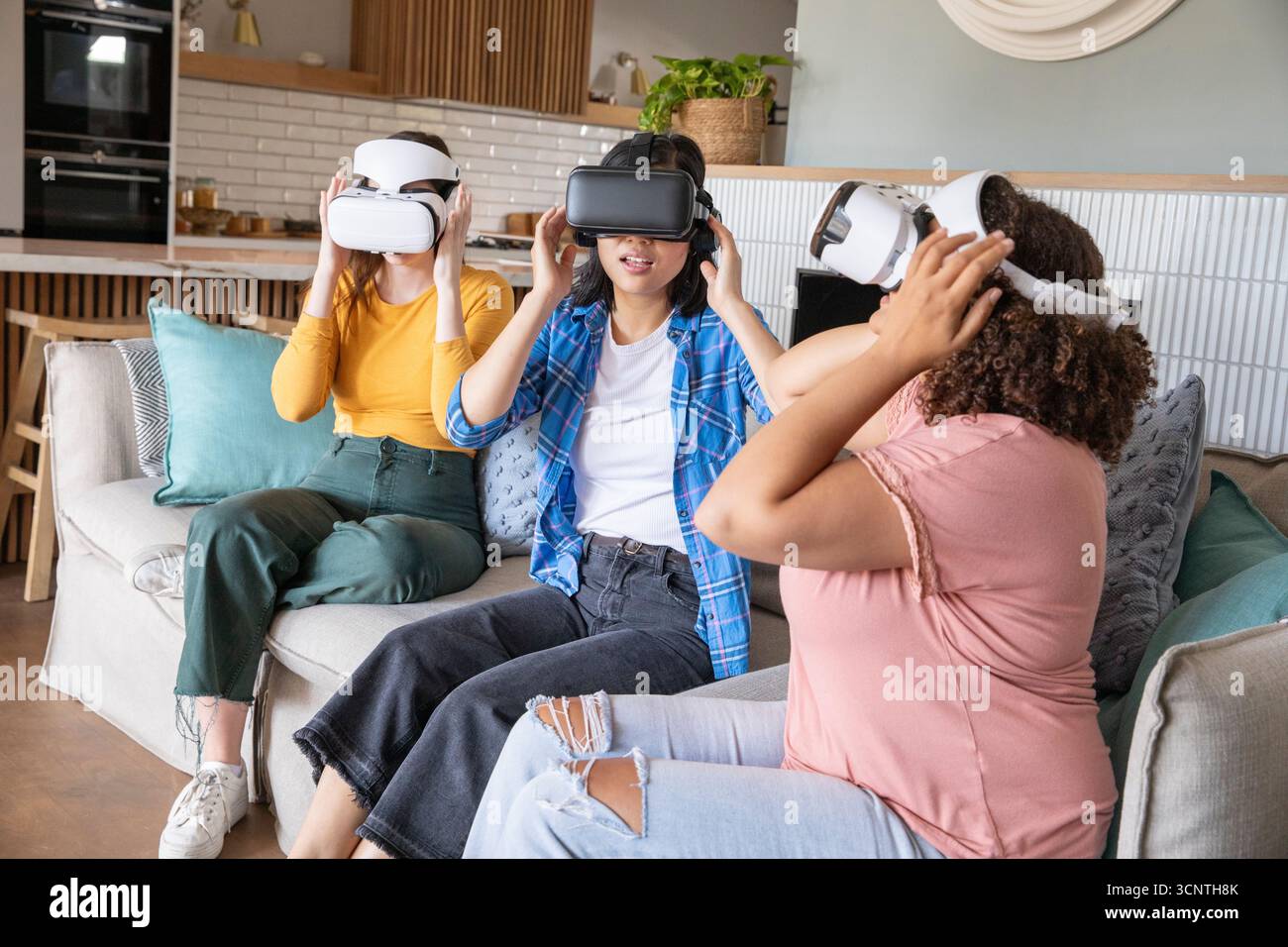 Verschiedene weibliche Freunde passen VR-Headsets an, die auf dem Sofa mit Kissen neben der Küche sitzen Stockfoto
