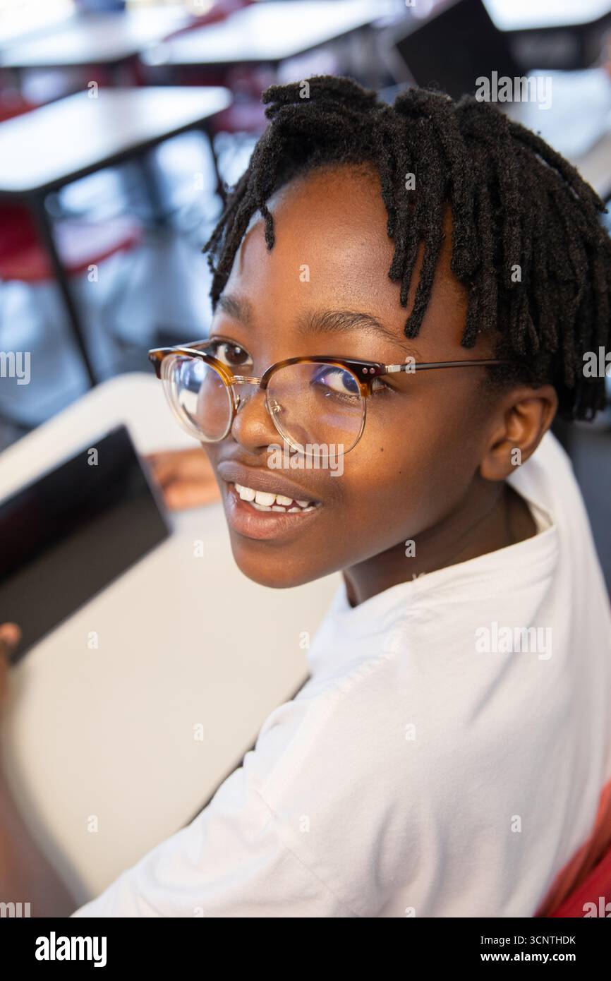 afroamerikanischer Junge mit Schildpatt-Brille und Tablet am Schreibtisch im Klassenzimmer mit Stühlen Stockfoto