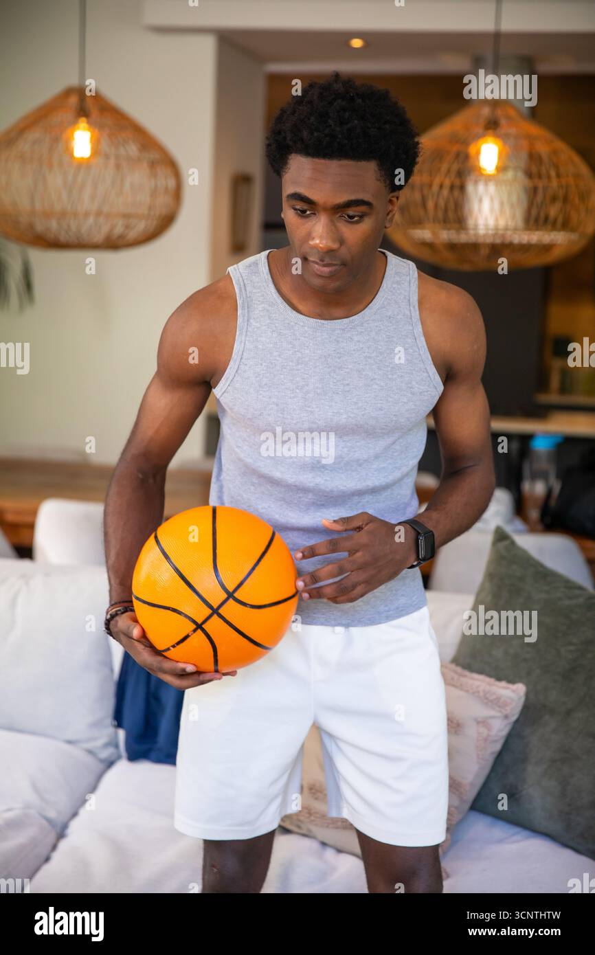 Afroamerikanischer Mann in Sportbekleidung, der Basketball in der Taille hält, während er im Wohnzimmer steht Stockfoto