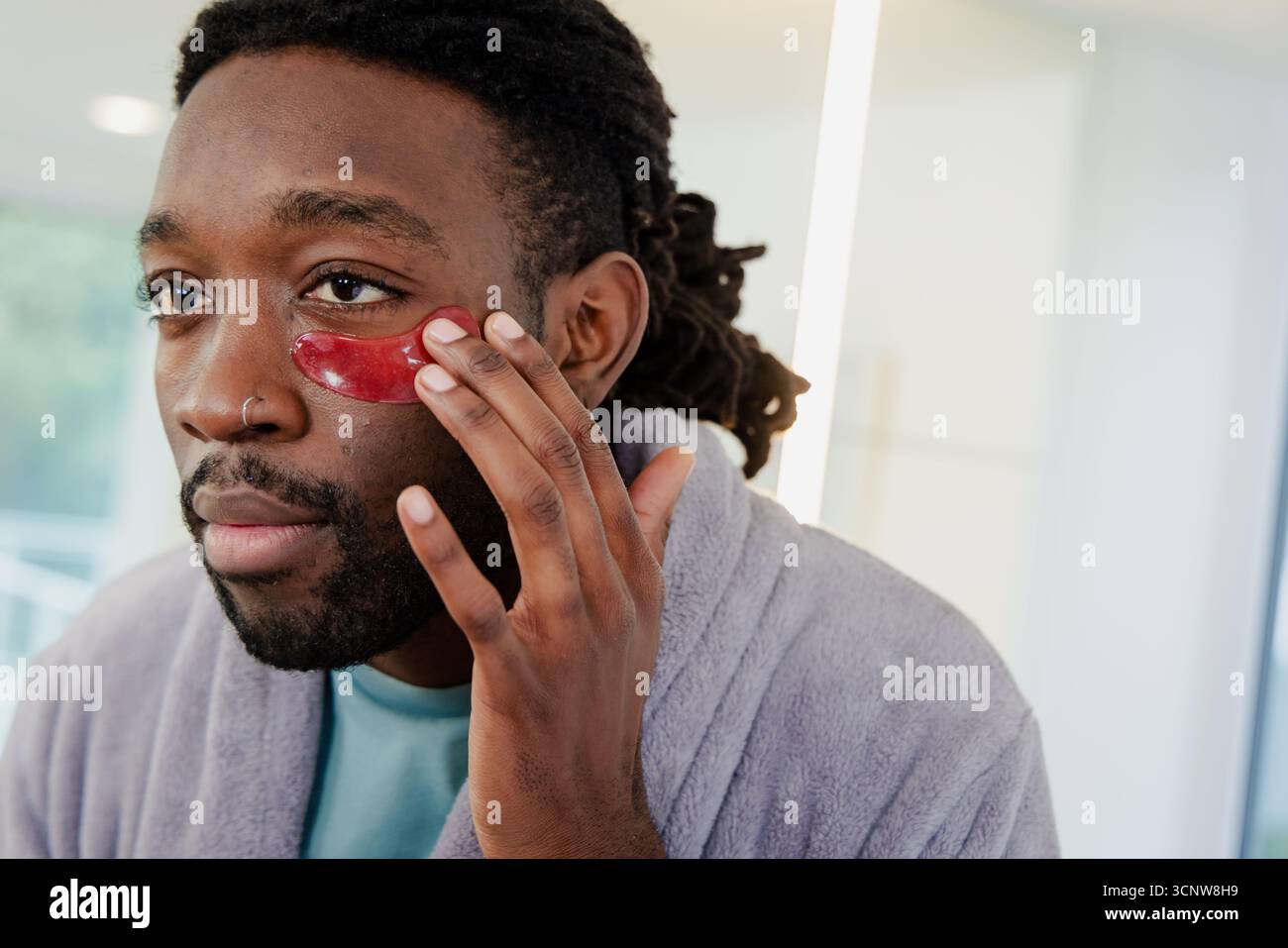 Afroamerikaner, der rote Hydrogel unter den Augen aufträgt und Bademantel im Badezimmer trägt Stockfoto