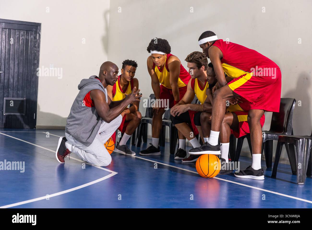 afroamerikanischer Trainer kniet auf dem Platz, verschiedene Spieler in roten gelben Trikots mit Ball Stockfoto