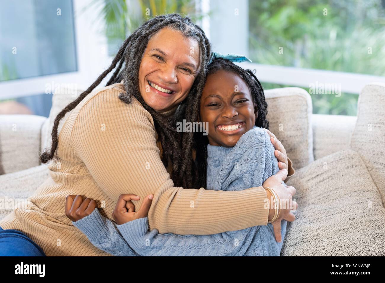Verschiedene ältere Großmutter und Enkelin, die sich an den Fenstern mit Kissen im Sonnenzimmer auf dem Sofa umarmten Stockfoto