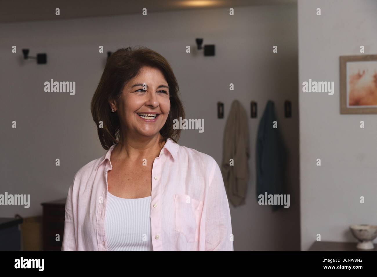 Frau, die im Flur zu Hause stand, trug ein hellrosa Hemd über weißem Tanktop zwischen Wandlampen Stockfoto