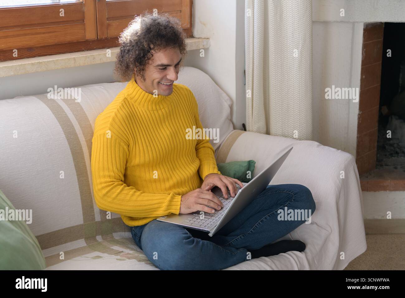 Ein Mann in einem gelben Pullover sitzt zu Hause bequem auf einem Sofa und benutzt einen Laptop. Er wirkt entspannt und konzentriert und genießt seine gemütliche, warme Umgebung Stockfoto