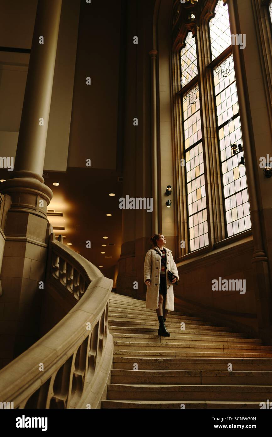 Eine Frau im langen Mantel steht auf einer eleganten Treppe in einem historischen Gebäude von Seattle und blickt auf große, verzierte Fenster, die den Raum füllen Stockfoto