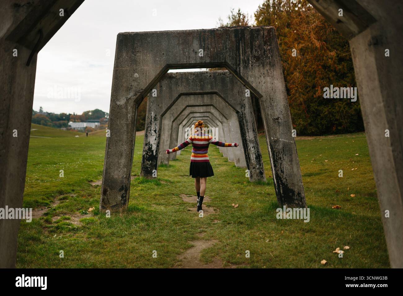 Eine junge Frau in einem farbenfrohen Pullover spaziert durch eine geometrische Architektur in einem Seattle Park. Umgeben von Herbstlaub, die Szene Captu Stockfoto