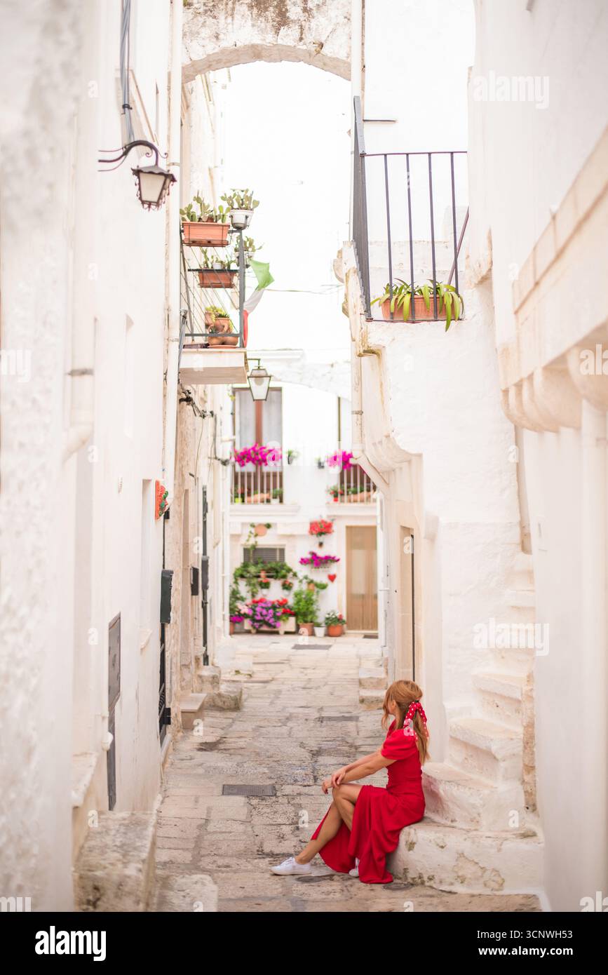 Eine Frau in rotem Kleid sitzt friedlich in einer malerischen Gasse von Cisternino, Apulien, umgeben von weiß getünchten Wänden und lebendigen Blumen, die das Land fängt Stockfoto