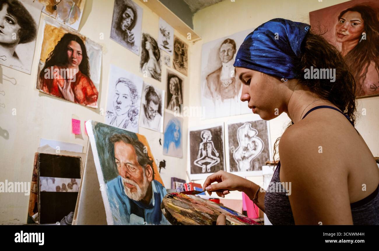 Eine junge Künstlerin konzentriert sich auf ihre Malerei in einem persönlichen Atelier, umgeben von expressiven Kunstwerken. Die Atmosphäre ist voll von Kreativität und Einflüssen Stockfoto