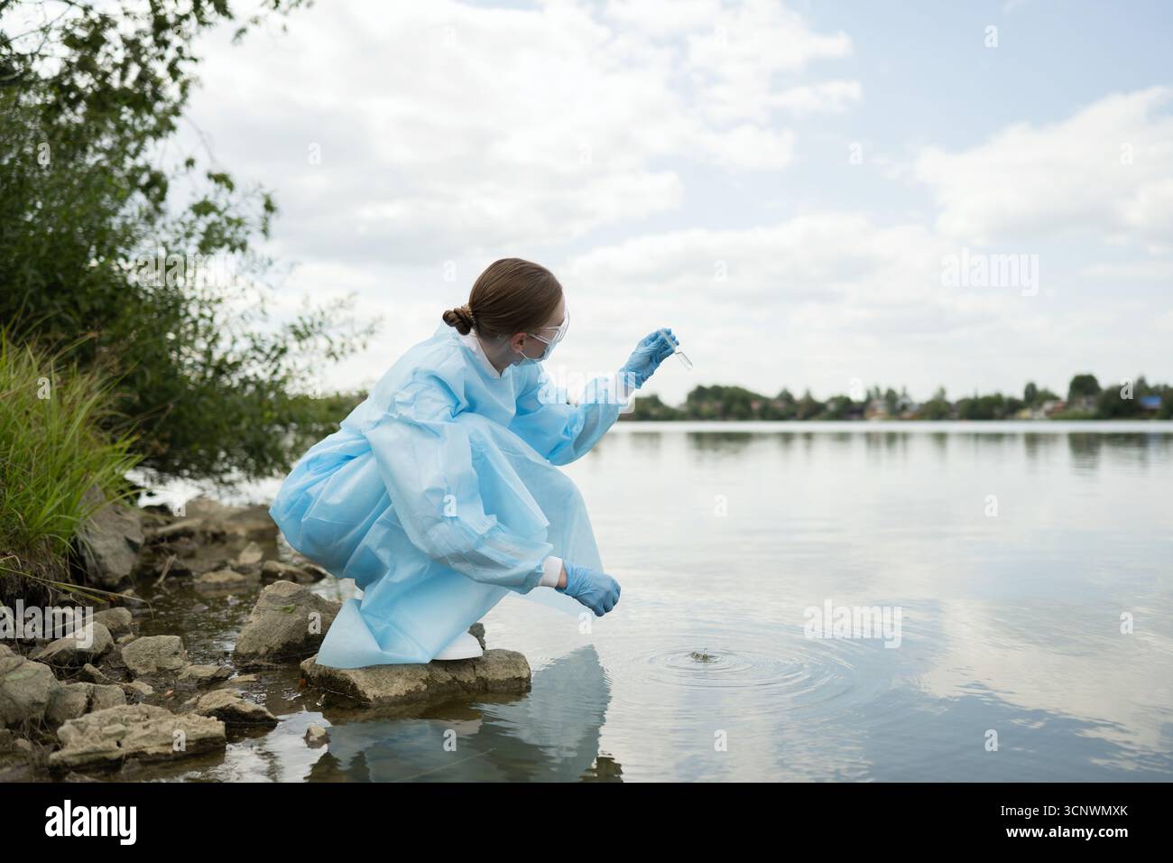 Eine junge Biologin in Schutzausrüstung führt Wassertests an einem ruhigen See durch und analysiert die ökologische Gesundheit. Die Einstellung betont die Verbindung Stockfoto