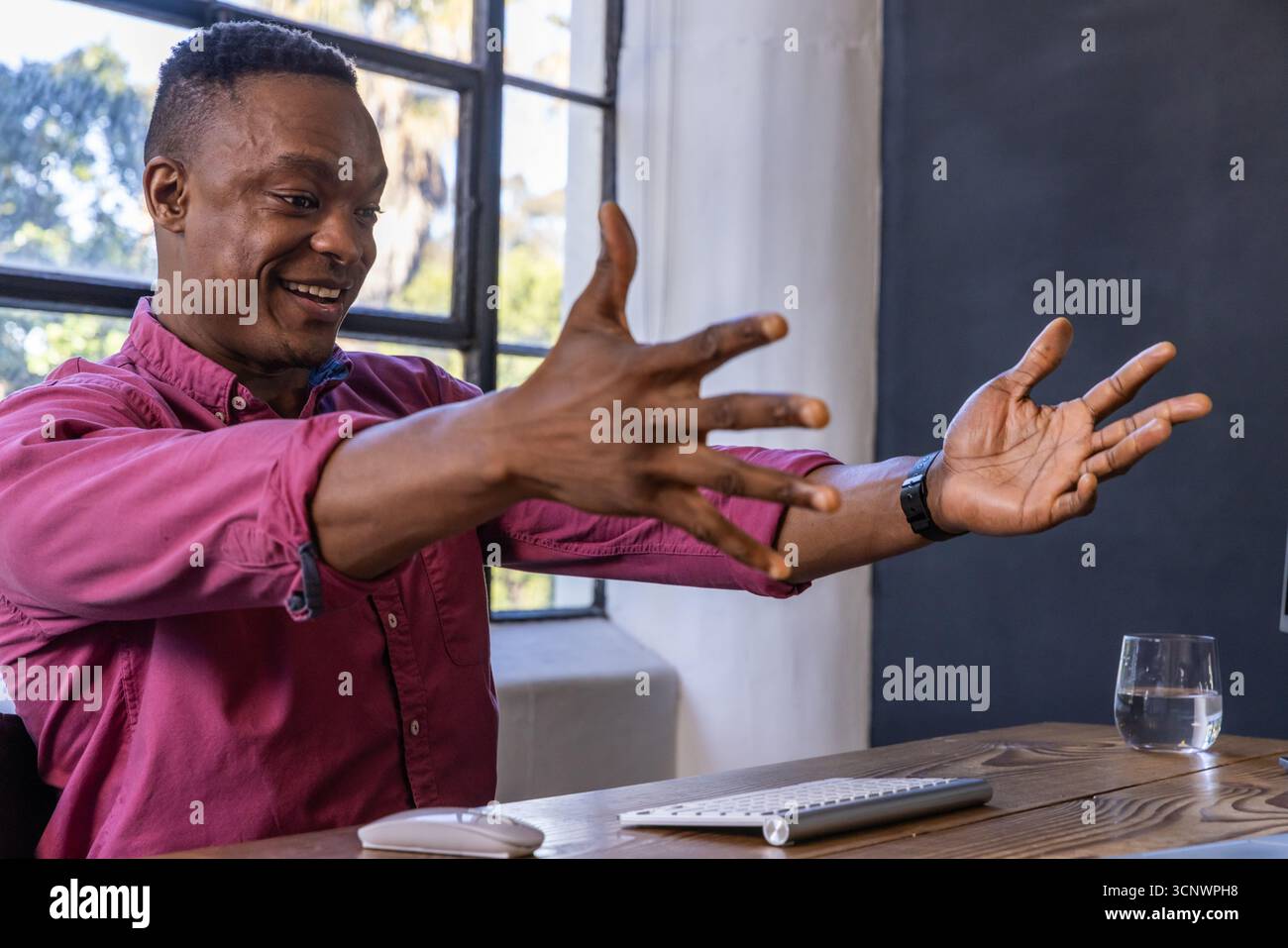 afroamerikanischer Mann, der am Schreibtisch Gesten mit Tastatur, Maus, Wasserglas macht Stockfoto