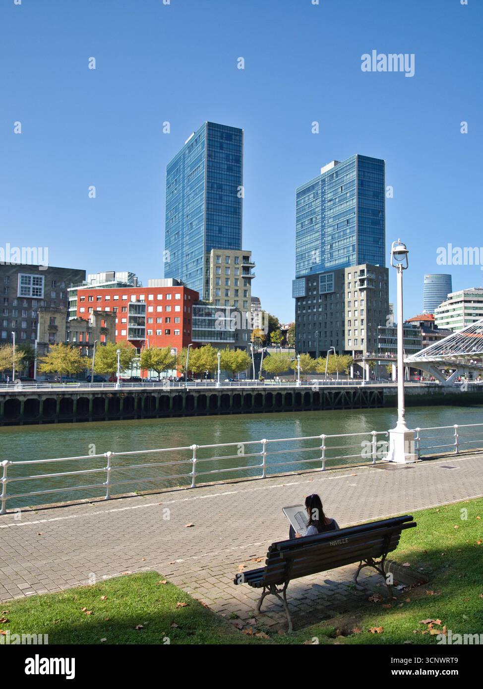 Friedliche Stadt Zuflucht am Fluss. - Eine junge Frau entspannt sich auf einer Bank, liest ein Buch, mit der modernen Skyline von Abandoibarra und dem Fluss Nervión Bilba Stockfoto