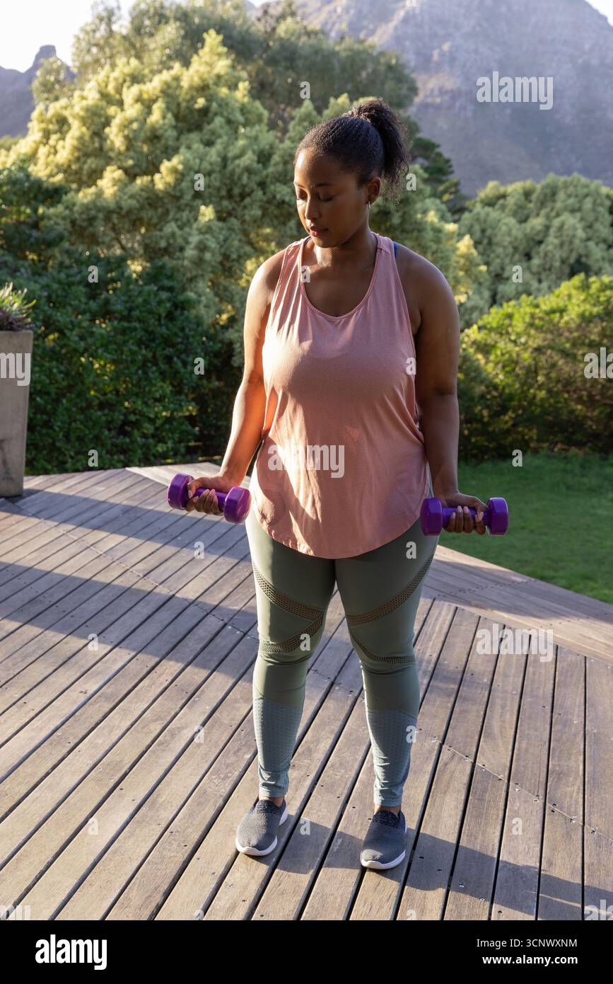 MittelErwachsene afroamerikanische Frau mit Kurzhanteln auf Deck mit Blick auf Garten und Berge Stockfoto