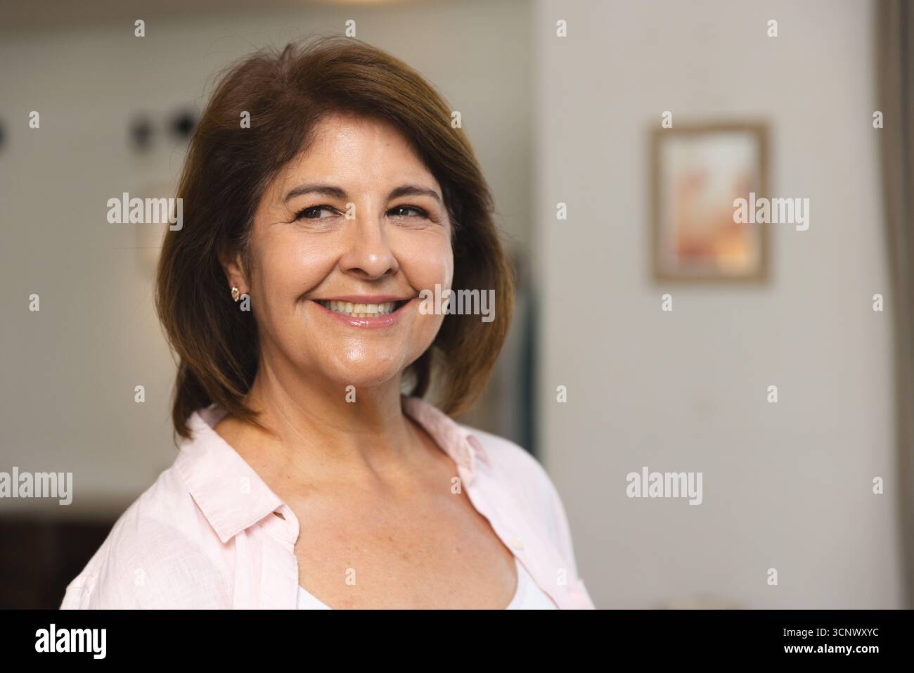 Frau lächelt, während sie eine hellrosafarbene Bluse über weißem Oberteil im Flur mit gerahmtem Bild trägt Stockfoto