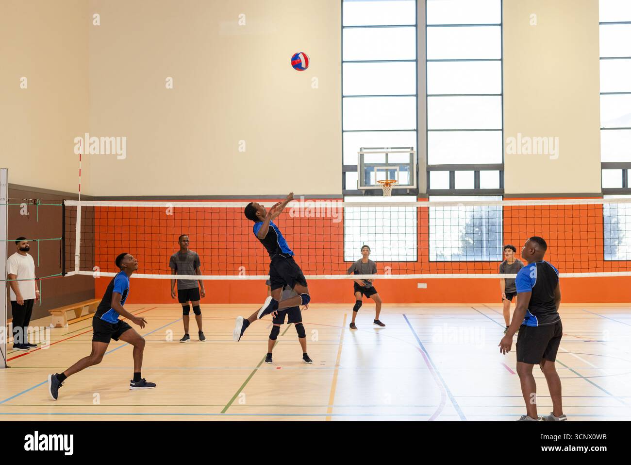 Asiatischer männlicher Volleyballspieler, der in der Nähe des Netzes springt und Volleyball im Fitnessstudio unter den Fenstern spielt Stockfoto