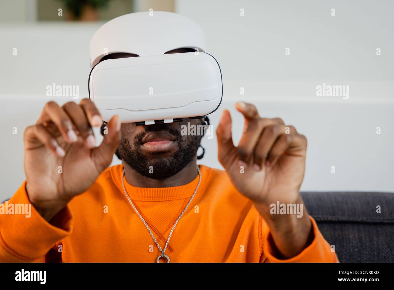 Das weiße VR-Headset liegt auf dem dunklen Sofa im modernen Wohnzimmer mit einem leuchtend orangefarbenen Sweatshirt Stockfoto