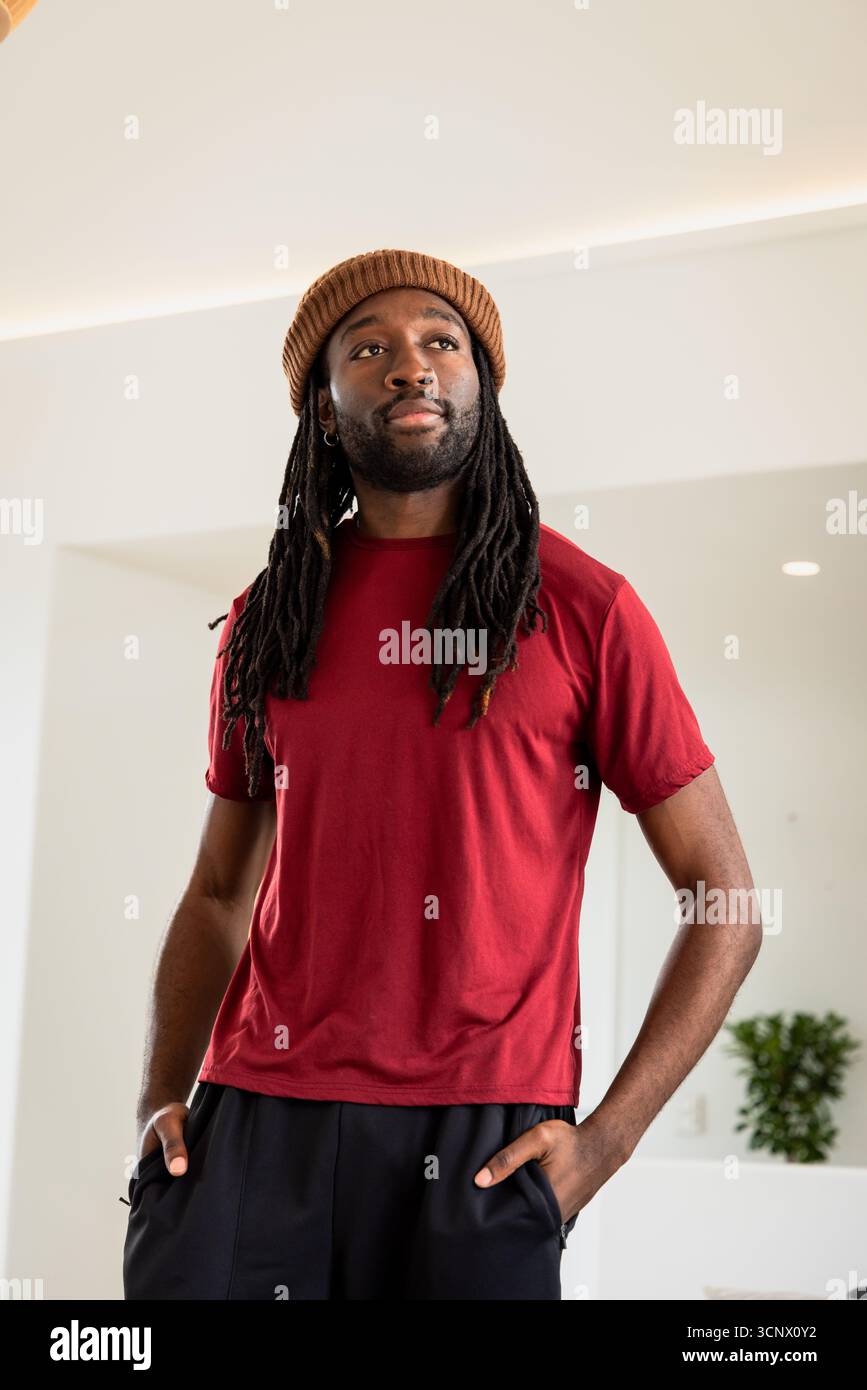 Afroamerikaner steht im Flur mit rotem Hemd, schwarzer Hose, brauner Beanie in der Nähe von Topfpflanzen Stockfoto