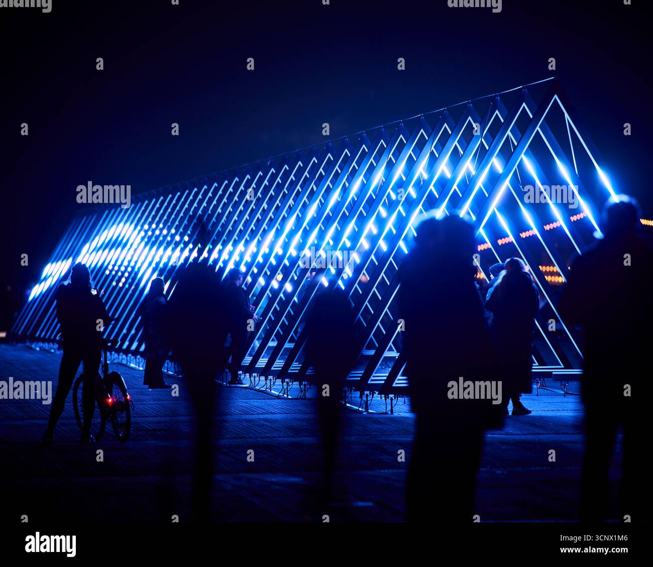 Blaue LED-Dreiecke bilden bei Nacht einen auffälligen geometrischen Tunnel, durch den Silhouettenfiguren laufen. Die Verschmelzung von Technologie und menschlicher Präsenz Stockfoto