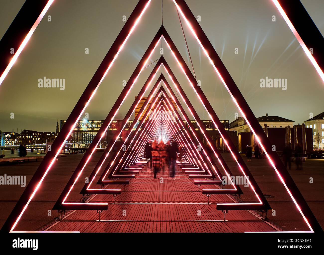 Rote LED-Dreiecke bilden bei Nacht einen auffälligen geometrischen Tunnel, durch den Silhouettenfiguren laufen. Die Verschmelzung von Technologie und menschlicher Präsenz Stockfoto