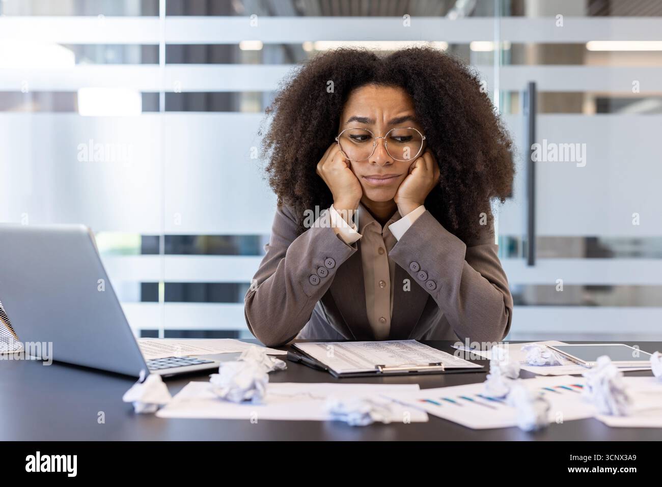 Müde junge afroamerikanische Frau, die am Schreibtisch im Büro sitzt und frustriert und verärgert über zerknittertes Papier, gescheiterte Arbeit und Ideen aussieht. Stockfoto