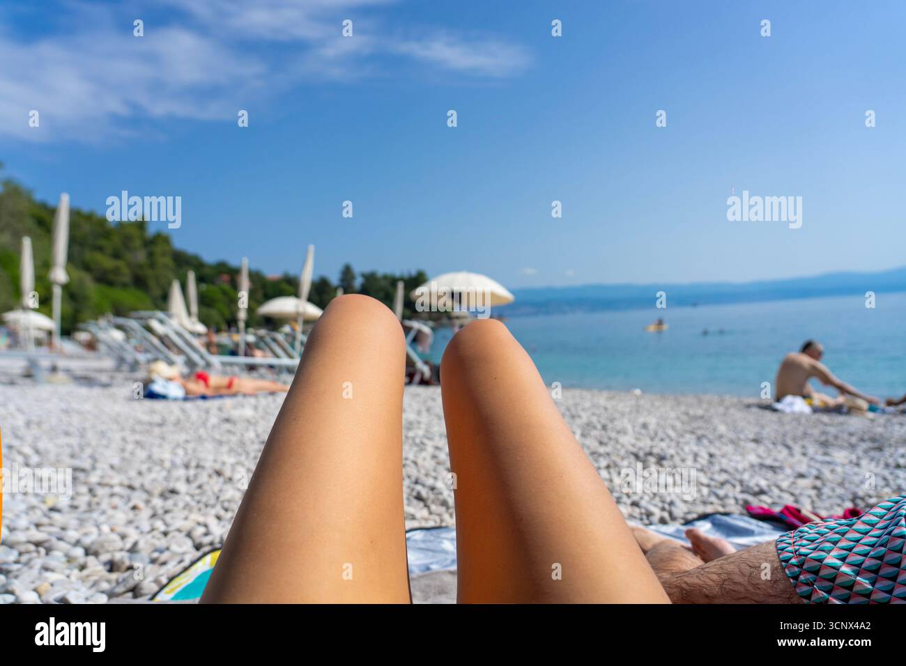 Entspannender Blick auf die gebräunten Beine an einem sonnigen Kiesstrand mit Sonnenschirmen und Meer am Medveja Strand Kroatien. Stockfoto