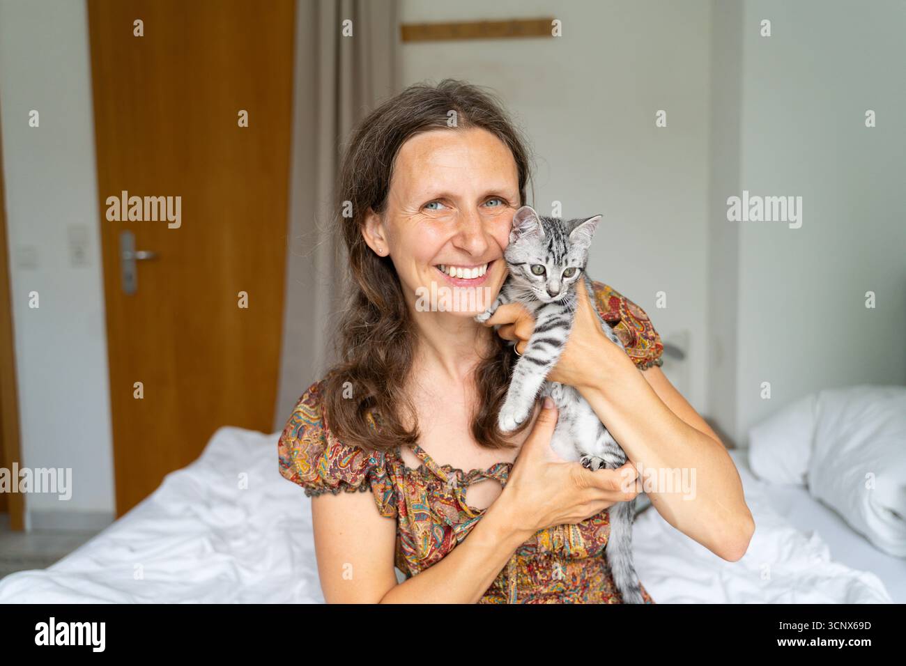 Eine Frau hält ein verspieltes graues Kätzchen in den Armen und lächelt warm, während sie auf einem Bett sitzt. Die gemütliche Wohnatmosphäre verleiht Ihrem Zuhause ein Gefühl von Komfort und Freude Stockfoto