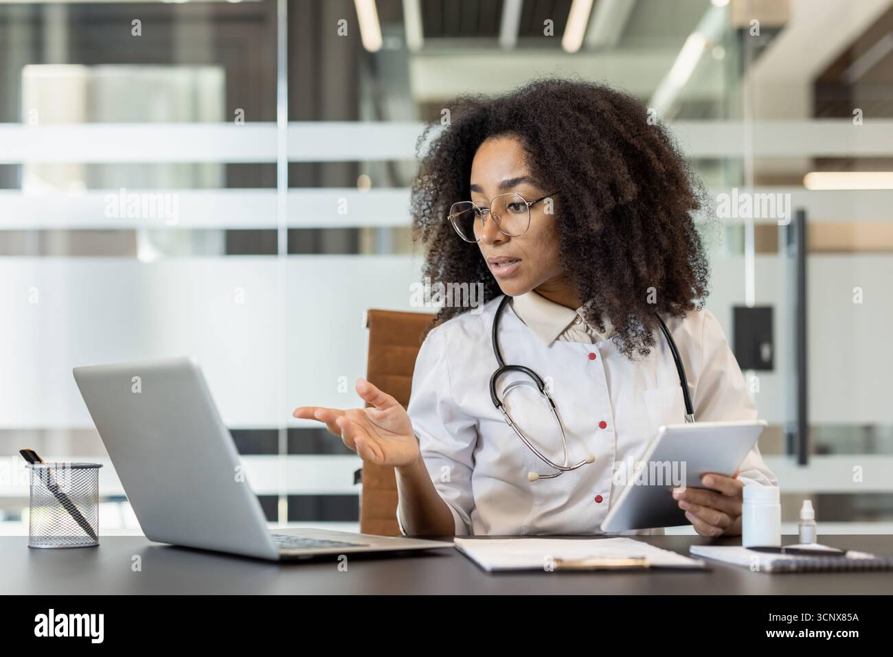 Konzentrierte und ernsthafte junge afro-amerikanische Ärztin sitzt im Klinikbüro am Tisch vor dem Laptop, hält das Tablet in der Hand und konsultiert den Patienten online, führt Videokonferenzen durch. Stockfoto